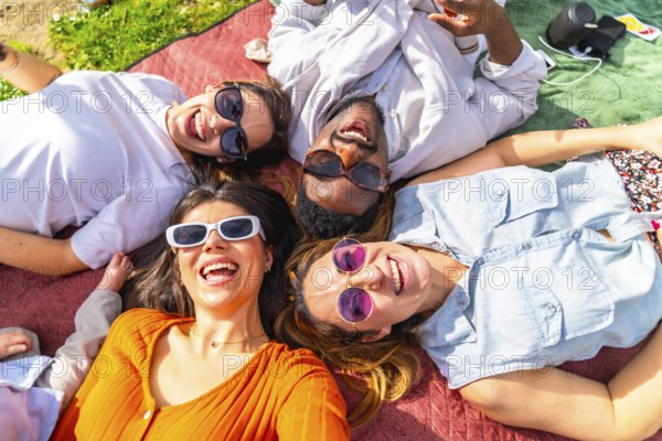 Four cheerful young adults lying together on blanket, enjoying sunny day picnic in park, listening to music and smiling