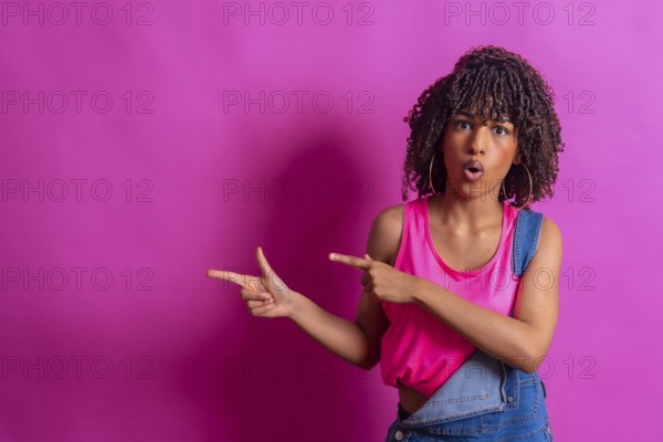 Surprised young woman with curly hair points to the side with a shocked expression, suggesting a promotional opportunity against a vibrant pink backdrop
