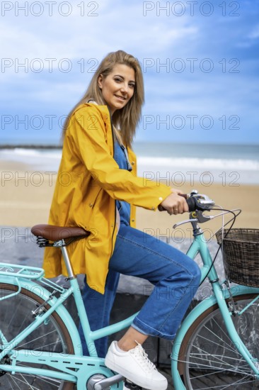 Young woman with yellow raincoat sitting on a turquoise vintage bicycle by the beach, enjoying a sunny day