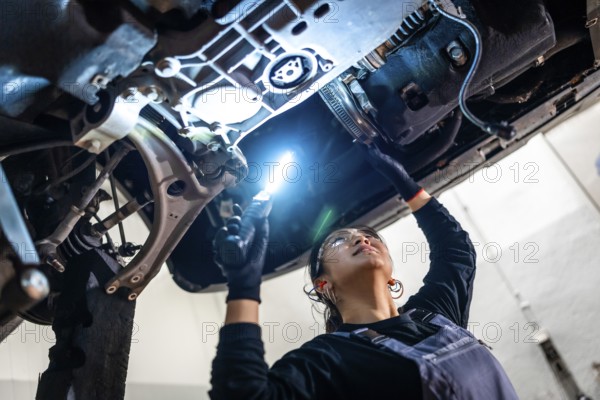 Female auto mechanic inspecting the underside of a car in a repair shop, using a flashlight to check for potential issues and ensure safety
