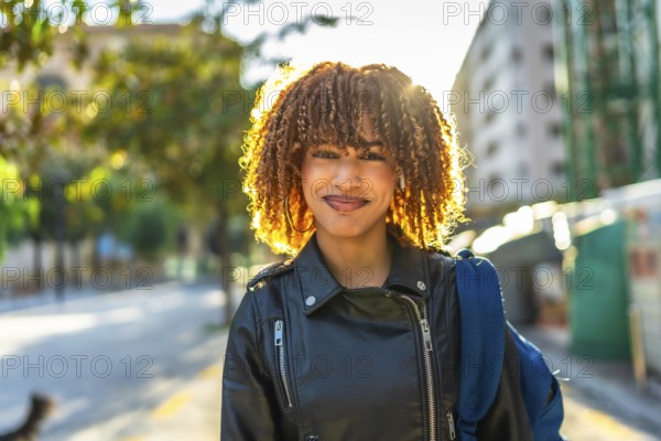 Confident young latin woman with curly hair and leather jacket smiling at camera standing in the street during sunset
