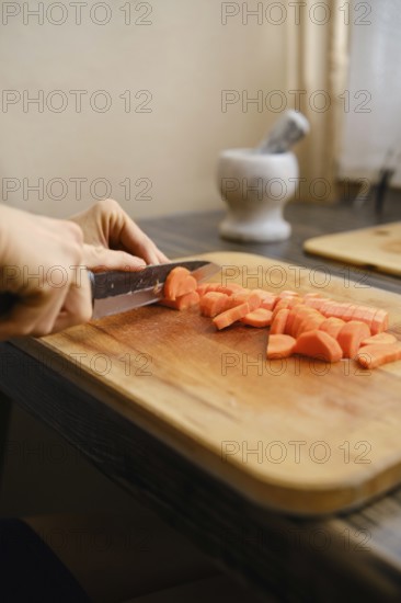A person skillfully slices fresh carrots into bite-sized pieces on a wooden cutting board. Sunlight filters through a nearby window, creating a warm and inviting atmosphere for cooking