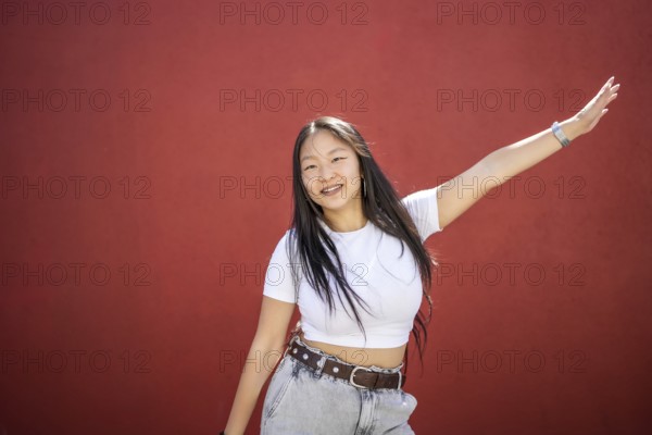Frontal portrait of a happy beauty young asian woman raising arm dancing K-pop against a red urban wall