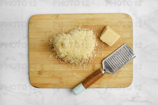 Shredded cheese on a wooden cutting board alongside a block of cheese and a grater. The items are arranged neatly in a well-lit kitchen, suggesting preparation for a delicious meal