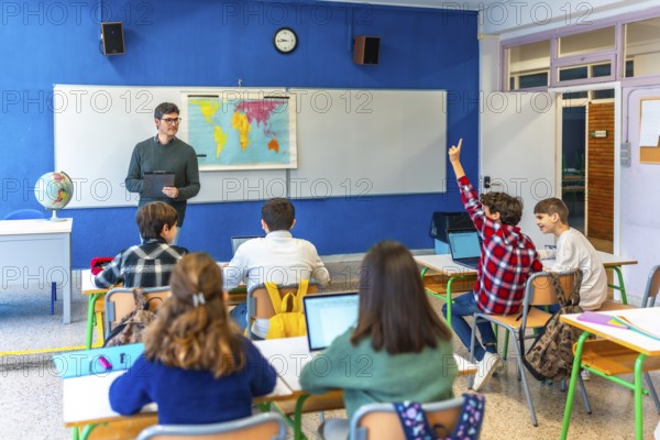 Group of teenage students raising hand and following geography lesson in classroom with teacher and laptops
