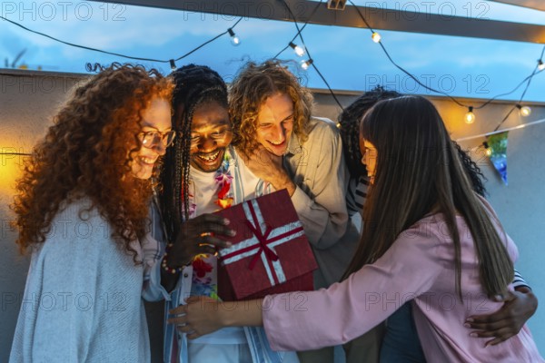 African man opening birthday present embraced by his multiracial friends during rooftop party