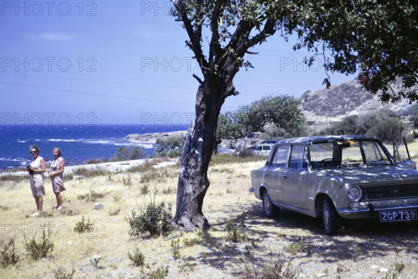 Two women standing by coast with Fiat 124 car parked in shade of olive tree, Mediterranean Sea, Cyprus, Europe 1973, Europe