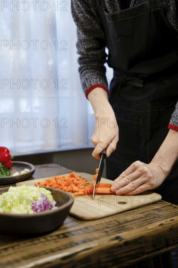 Unrecognizable man is chopping vibrant carrots on a wooden cutting board while surrounded by fresh vegetables like bell peppers and celery. Bright light filters through the window, creating a warm atmosphere.
