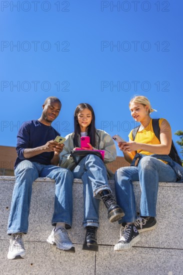 Vertical low angle view photo with copy space against blue sky of multi-ethnic students sitting on a wall outside the campus using mobile phones