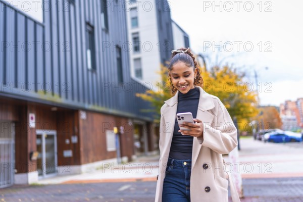 Happy stylish african american young woman using phone walking in the city in autumn
