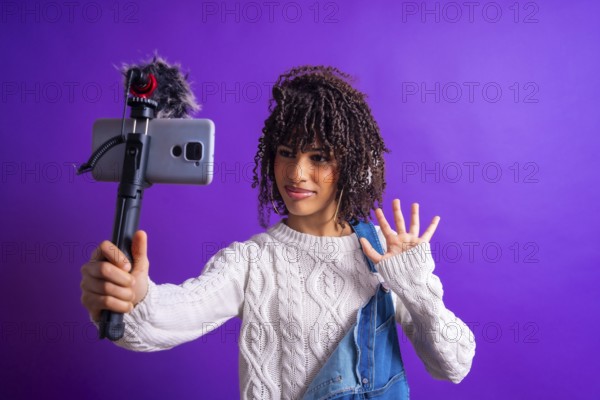 Young influencer waving while recording a video blog using a smartphone with external microphone on purple background