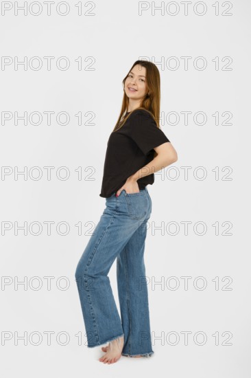 Barefoot woman stands with a playful posture, smiling at the camera while wearing a black top and wide-leg jeans. The simple white background emphasizes her casual, confident demeanor and style