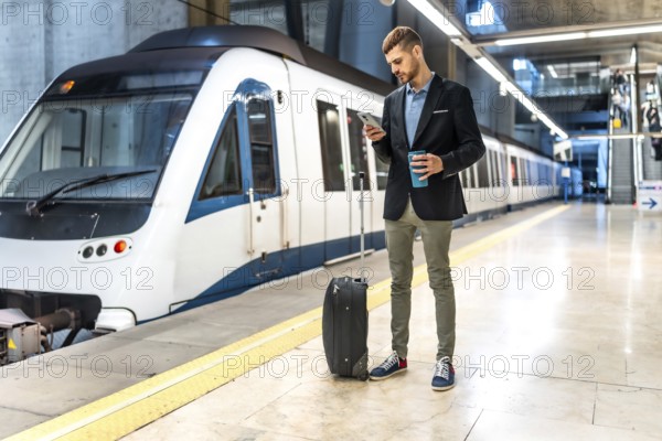 Young businessman standing on subway platform with suitcase, using smartphone and holding reusable coffee cup while waiting for train