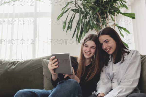 Two teenager sister girls are sitting at green sofa in living room and watching some funny video at tablet, smiling, laughing. Beautiful people, frendship and togetherness concept. Flowerpot on back