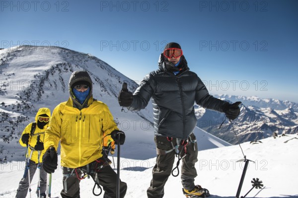 A middle-aged climber in a down jacket and harness shows a thumb up next to his friends on the way to the top of a snow-capped mountain. The concept of rest and climbing high mountains with friends