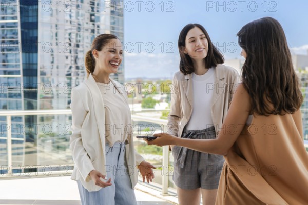 Three women are standing on a rooftop, smiling and talking to each other. One of them is holding a cell phone