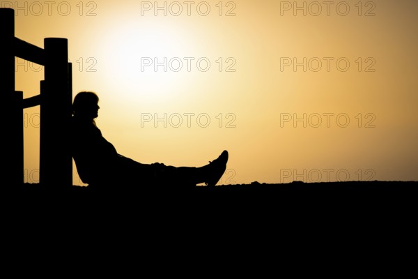 Silhouette of an unrecognizable person sitting against wooden pole with orange colored sunset in the background