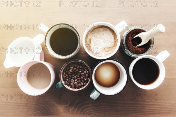 Multiple coffee cups and multicolored mugs with americano, espresso, latte and cappuccino, milk jar, roasted coffee beans in glass bottle, wooden spoon and ground coffee in jar on wooden background