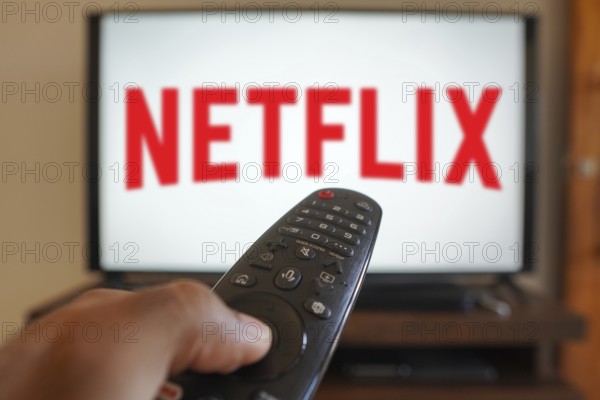 April 20, 2024, Brazil. In this photo illustration, a hand holding a TV remote control in front of the Netflix logo on a TV screen