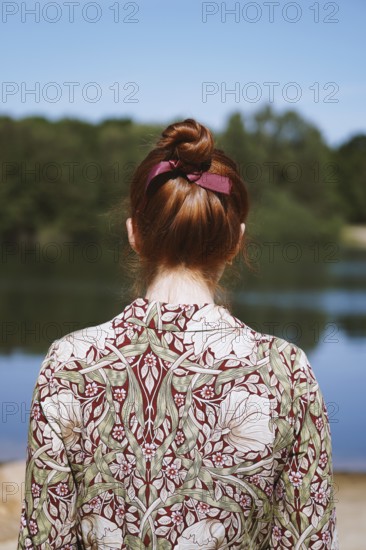rear view of anonymous young woman with floral dress and red hair bun standing by lake in solitude or depression