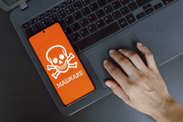 In this photo illustration, the Malware icon is displayed on a smartphone screen next to a person using laptop