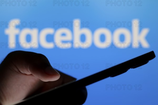 In this photo illustration, the Facebook logo seen in the background of a silhouette people holding a smartphone