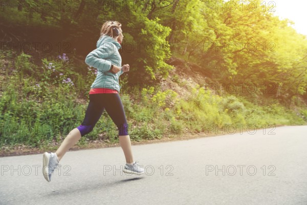A young blonde woman running is practicing outdoors in a city mountain park in the forest. Warm rays through the branches of trees