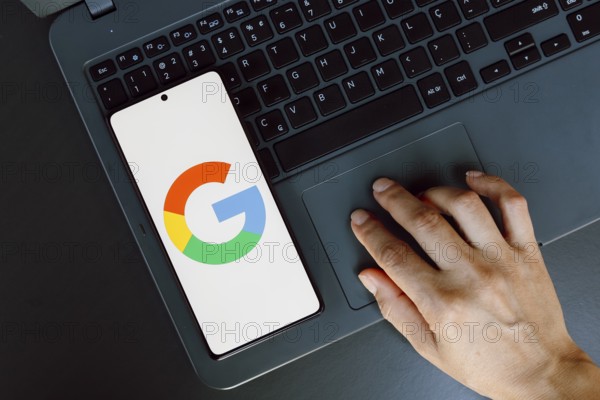 In this photo illustration, the Google logo is displayed on a smartphone screen next to a person using laptop