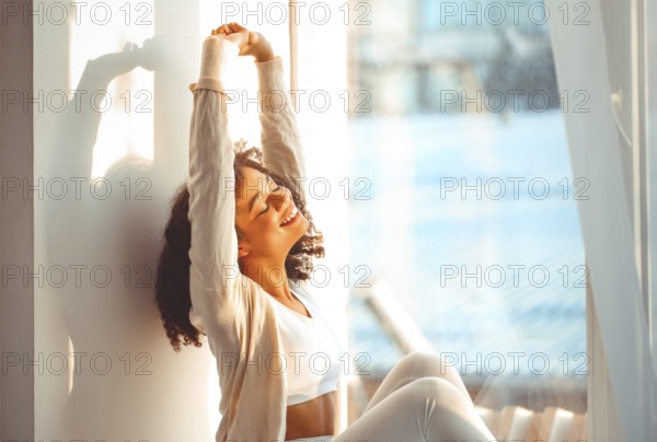 Pleased relaxed African American ethnicity girl in homewear sitting on windowsill against backdrop of curtains fluttering from light wind, happy afro female at home smiling gently with eyes closed
