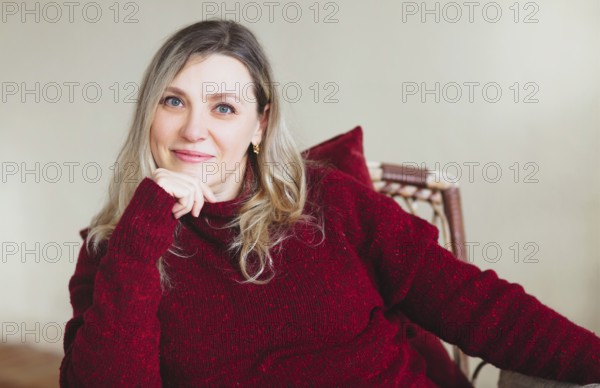Attractive middle aged happy woman sitting and relaxing on comfortable chair at home on weekend, wearing colorful knitted warm red sweater, smiling at camera. People lifestyle lifestyle