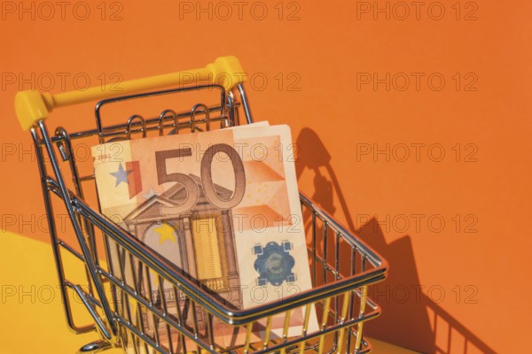 Euro paper currency 50 banknote money Shopping trolley cart on colorful orange yellow background. Copy space for your text. Online shopping, buy mall market shop consumer concept. Small toy supermarket grocery push cart. Food crisis. Rising food cost, grocery prices shortage mass hunger. Inflation
