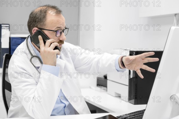 Male doctor pointing with finger on desktop computer while talking on the phone, discussing treatment with colleague. High quality photo