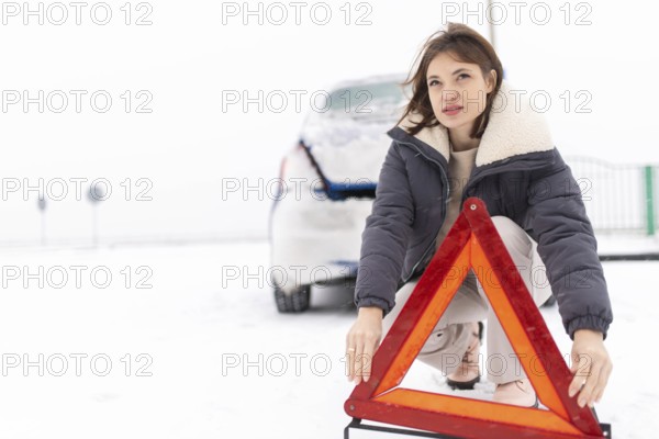 Woman puts up a warning triangle on a snowy road in winter, car breakdown in winter