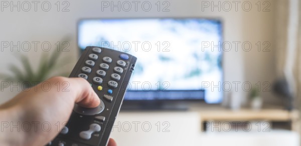 Holding a TV remote control in the hand, foreground, tv in the blurry background. Streaming