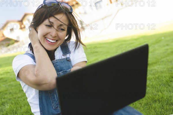 Female gets joyful emotions by talking to her friends on a conference call from notebook, she dressed in jeans overalls sitting on grass in the park