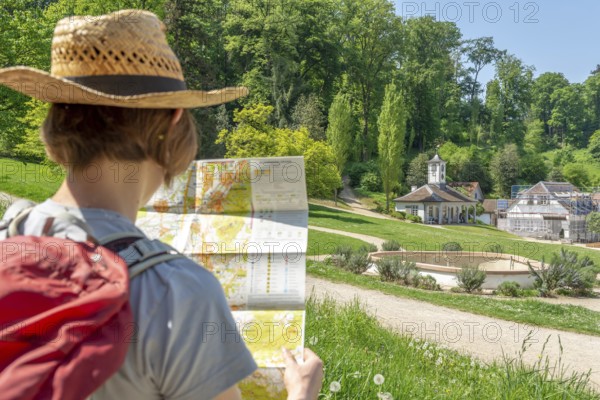 Woman with brown hair, gray t-shirt, jeans, red backpack and straw hat taking a break from her hike, sitting and looking on a hiking map, Staatspark Fürstenlager, Auerbach, Bensheim, Germany, Europe
