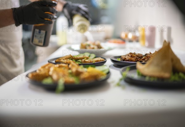 Chef wearing black gloves is pouring a sauce from a bottle on several plates of indian food, including samosas and pakoras, arranged on a white tablecloth in a restaurant kitchen
