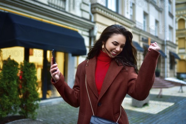 Beautiful Overjoyed young brunette woman with long hair in a red coat celebrating good news holding phone and clenching fist in the evening street