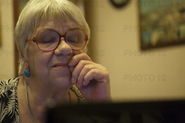 Thoughtful senior woman with blond hair wearing spectacles reading a computer screen with her hand to her face and an expression of concentration