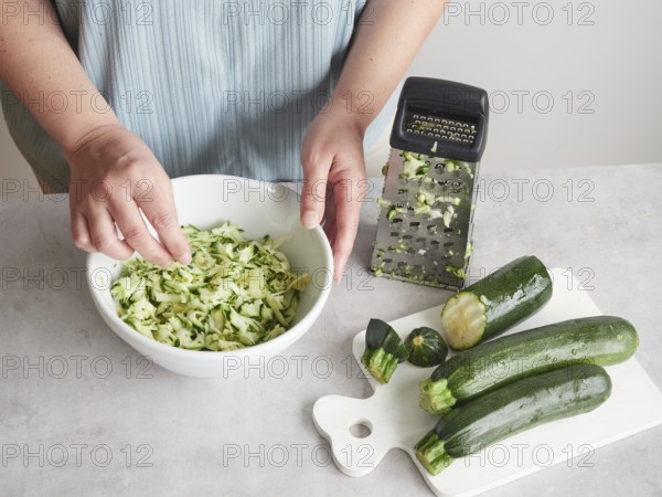 Woman with her hands in a bowl of grated zucchini, other fresh zucchini on wooden board and stainless steel hand grater on the countertop