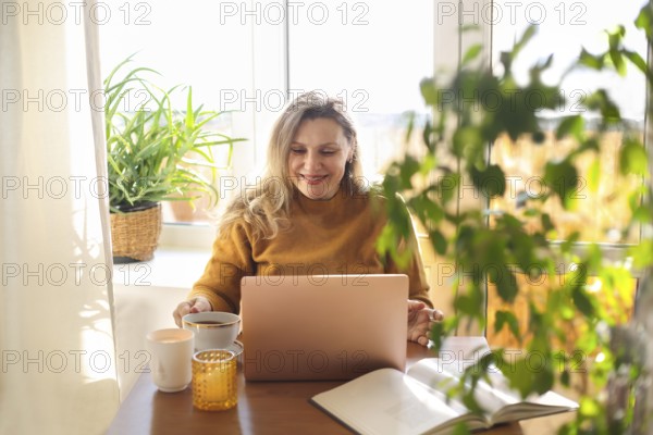 Joyful happy middle-aged woman chatting online on laptop at home, making video call via internet, overjoyed 50s female looking at computer screen spreading arms with happiness while sitting at table