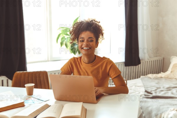 Cheerful young African American female student sitting at desk at home surrounded by textbooks, in front of laptop studying hard prepares for exams online, looking in camera with satisfied smile