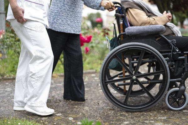 Senior woman in wheel chair with relative and nurse caregiver, outdoors at nursing home garden. graphy