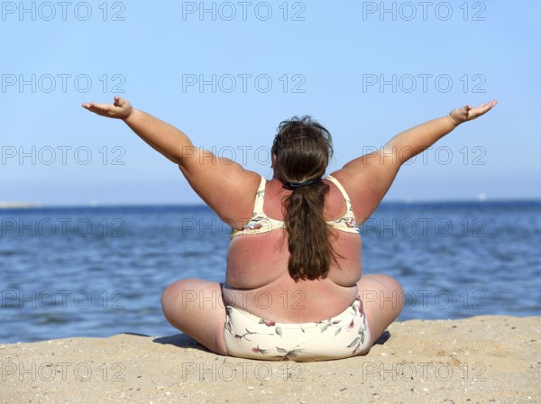 Overweight woman sitting on beach with hands up