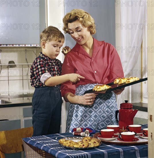 Baking in the 1950s. A mother with her son in the kitchen. She has freshly baked buns and cakes from the oven and the boy looks as if he likes the nice smell from them. A typical 1950s picture with the mothers clothes, the cups and can, the kitchen interior and the colors. Sweden 1958 Conard