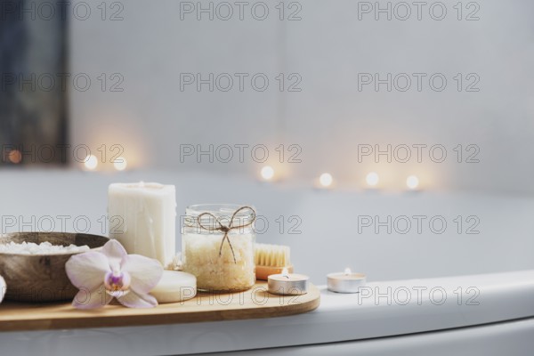Preparation for hotel spa treatment or home bath procedure. White washbasin in bathroom with accessories on tray. Burning candles, washcloth, soap, foot brush, bottle with sea salt, orchid flower