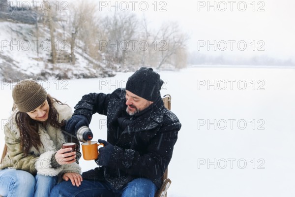 Happy lovely middle-aged couple in warm clothes drinking hot tea during romantic winter date in nature, man and woman in love sitting on frozen lake under falling snow and chatting on cold frosty day