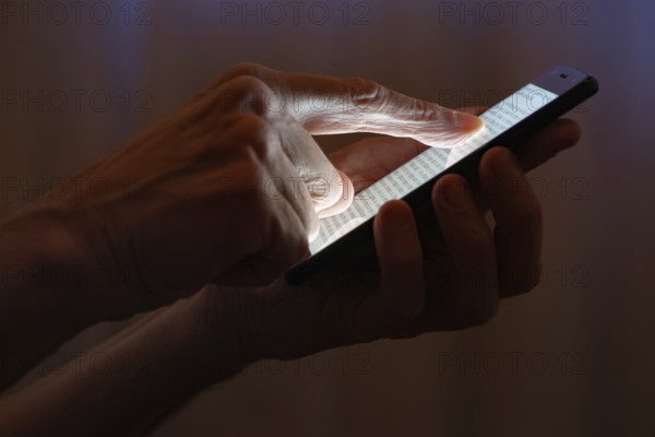 Man typing with the finger on the touch screen of a black smartphone close to the window with closed curtains at night
