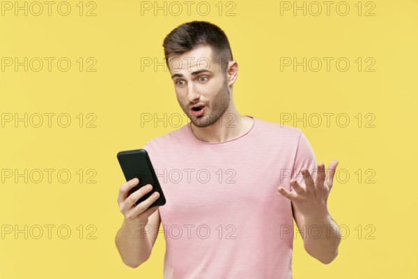 Shocked funny millennial man using mobile phone holding it in hand isolated over yellow background