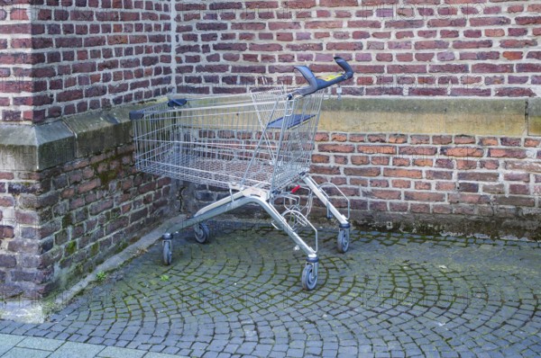 Metal shopping cart stands empty next to a brick wall on a cobblestone street, symbolizing consumerism, retail, and urban life, Bottrop, NRW, Germany, Europe
